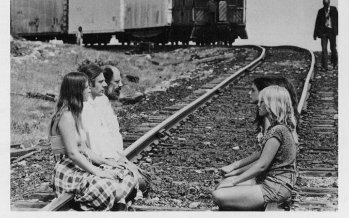 Auf einem Bahngleis sitzen sich Menschen gegenüber. Von hinten naht ein Zug.
