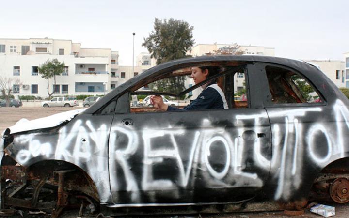 Eine Frau sitzt in einem funktionsuntüchtigen Auto. Auf der Fahrerseite wurde mit weißer Farbe »Revolution« gesprüht.