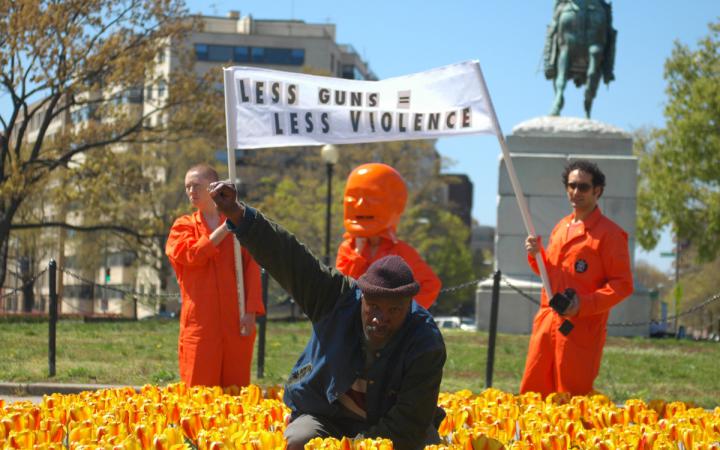 Ein Mann im Blumenbeet hebt seine rechte Faust als Zeichen des Protests. Hinter ihm drei Männer mit einem Transparent.
