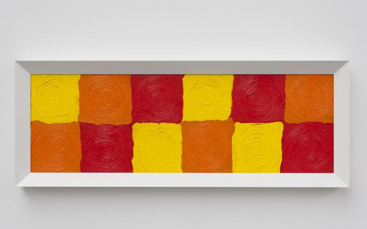 Zwölf farbig gemalte Quadrate in zwei Reihen übereinander in den Farben Gelb, Orange und Rot. Als Bild hängt es an der Wand und ist weiß gerahmt.