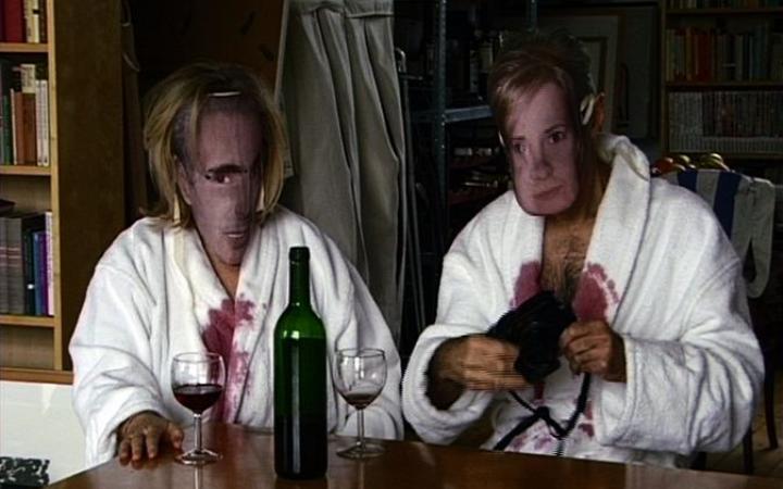 Zwei Personen mit Masken trinken Rotwein. Ihre Bademäntel sind dabei voller Rotweinflecken.