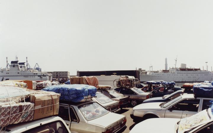 Fotografie mehrerer geparkter Autos, alle voll beladen. Im Bildhintergrund sind zwei große Kreuzfahrtschiffe.