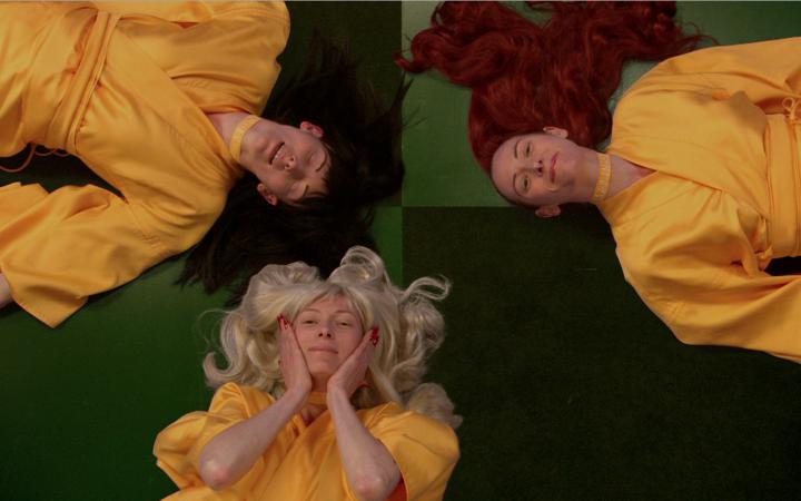 three women, each with blond, black and red hair color lie with their heads together on the floor and looking into the camera