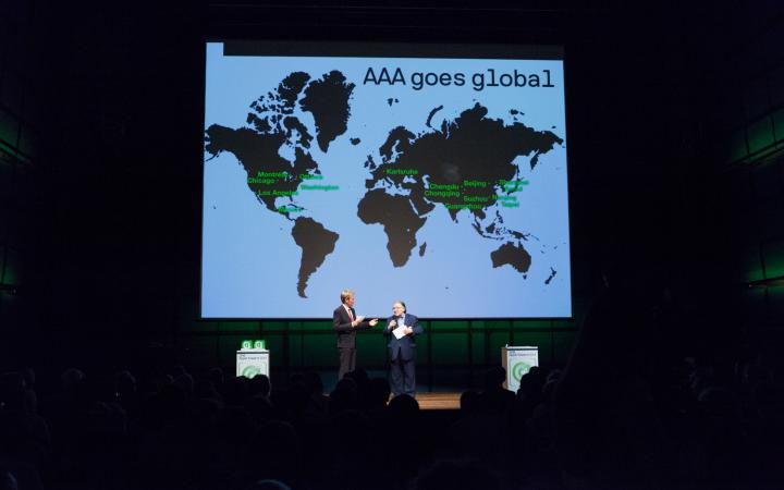 Zwei Männer auf der Bühne. Im Hintergrund eine Weltkarte mit »AAA goes global«