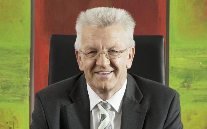 A laughing elderly gentleman. He is wearing a gray suit and a white shirt and a pair of glasses. His hair is gray. He sits on a black office chair. In the background, the colors are red and green to be seen.