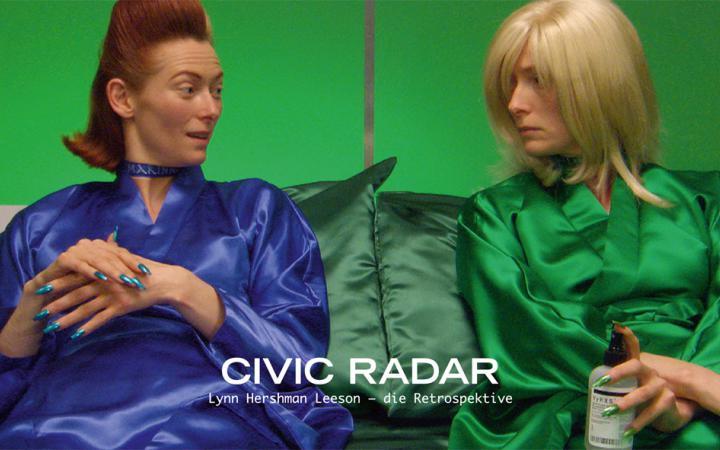 A woman in two different appearance. On the left side in the blue silk coat and brown hair, on the right side in the green silk coat and blond hair. Both sit together and look at each other.