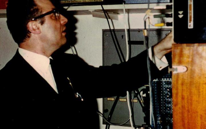 A man in a black suit and black horn-rimmed glasses served various electronic equipment.