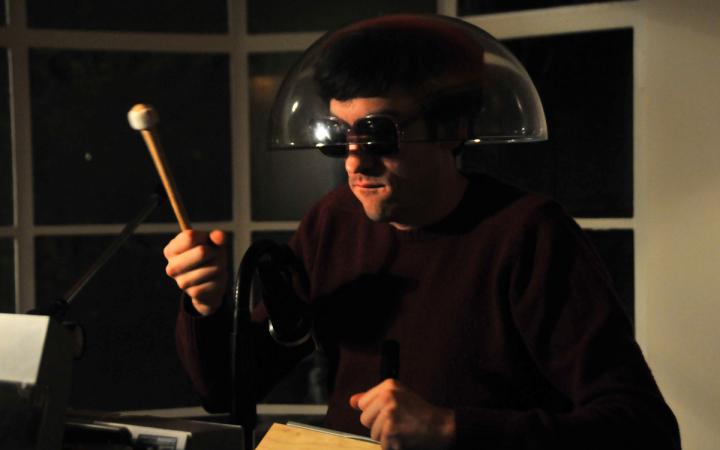 A man wearing sunglasses and transparent lampshade on his head, holding a drumstick in his right hand.