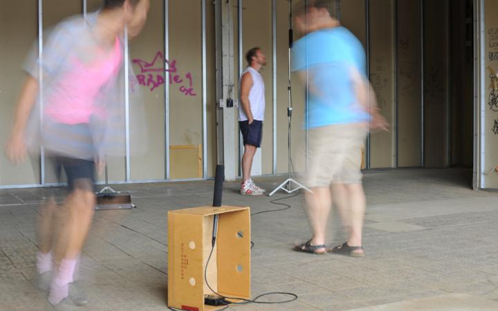 A banana box into which a microphone was installed. To the crate running around two men. By distorted blur they are not visible. In the background: Another person in front of a microphone.