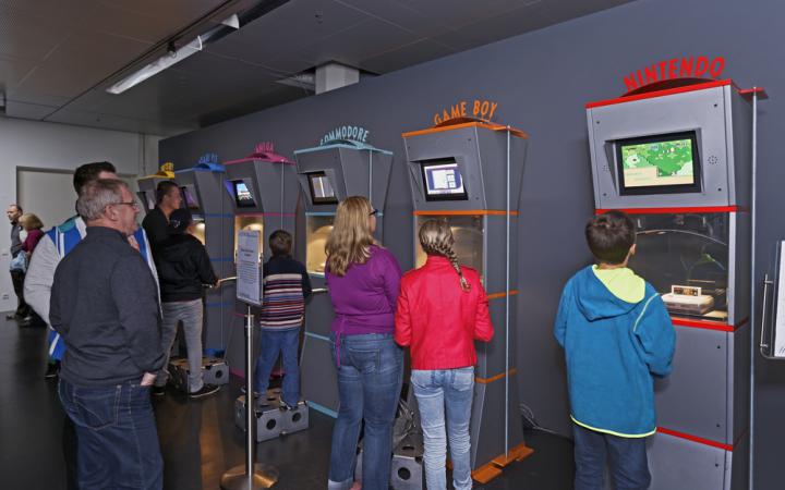Several people stand in front of 6 terminals and play computer games