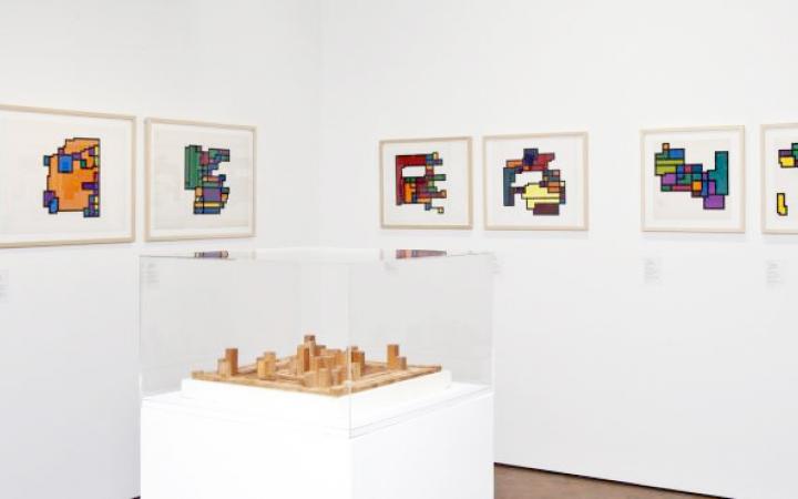 A view of the exhibition »Hiroshi Kawano«: A room with seven paintings with geometric patterns, in the middle an abstract wooden sculpture.