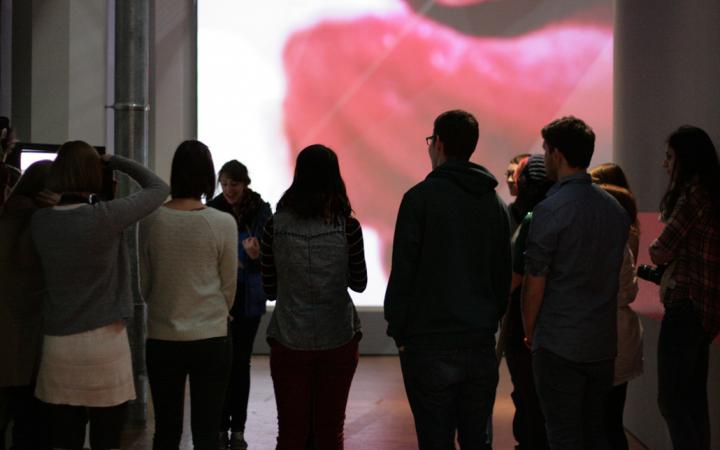 Eine Gruppe von Menschen vor einer Kunstvermittlerin. Im Hintergrund ist großflächig ein Teil einer Zunge zu sehen.