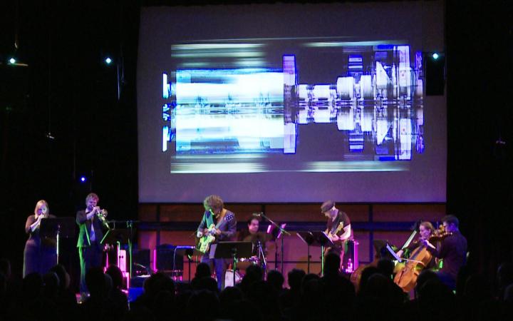 Musizierende Band auf einer Bühne. Über ihnen im Hintergrund futuristische Videoanimationen.