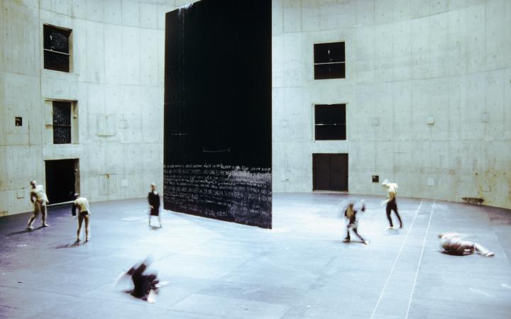Seven dancers move around a large rectangular black wall that is structured a theatrical space.