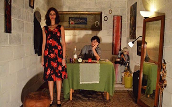 Blick in Zimmer mit Spiegel und Tisch. Dieser hat eine grüne Tischdecke. An dem Tisch sitzt ein Mann. Daneben steht eine Frau in einem schwarz rot geblümten Kleid.