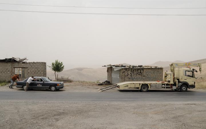 Zwei Männer schieben einen Mercedes in Richtung eines Abschleppautos. Hinter ihnen ruinöse Gebäude und eine karge hügelige Landschaft.