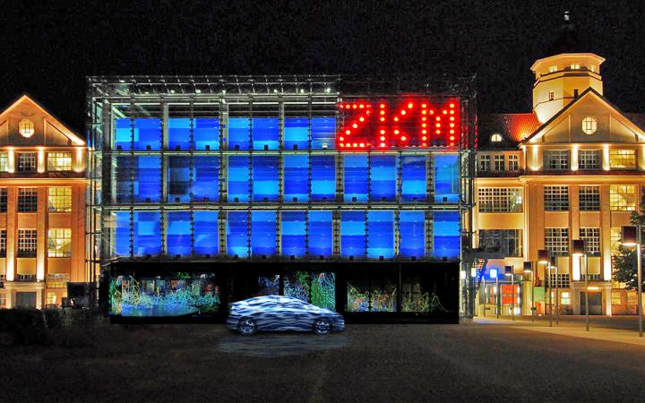 A car stands in front of the blue cube. Top right in big red glowing letters: ZKM