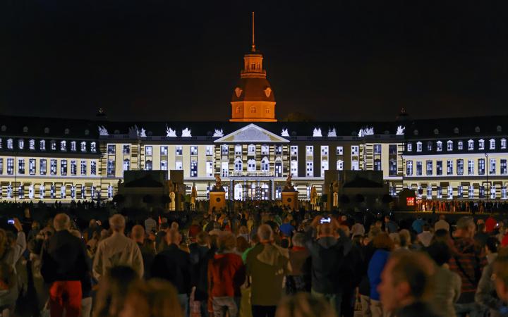 People standing in fron of the Karlsruhe palace, on which figures are projected