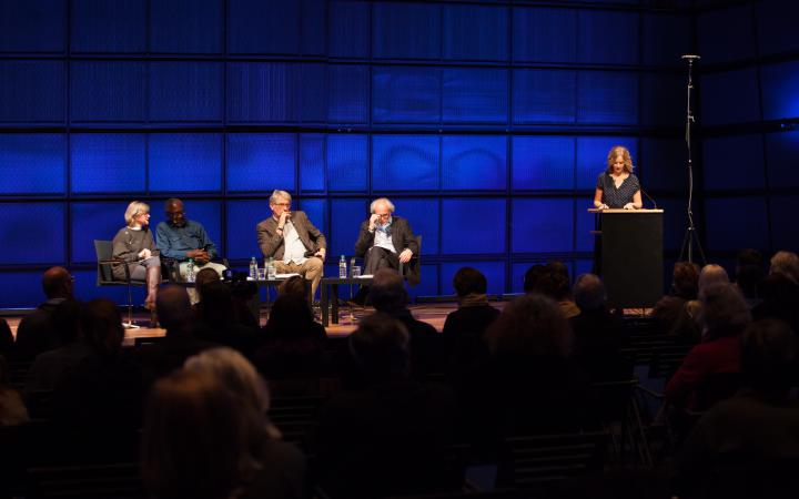 Four people sitting on a stage. Another person stands right by them on a pedestal and speak to the audience