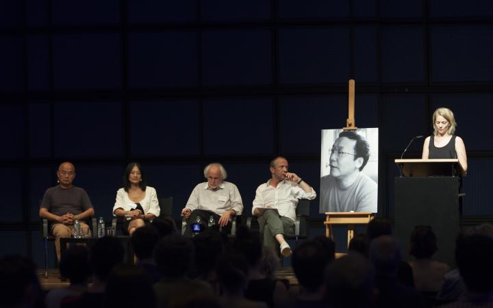Four people sitting on the stage. At the lectern talking woman and a large black and white photo of a young man is placed.