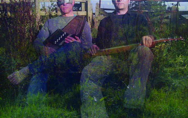 Zwei Männer sitzen mit Musikinstrumenten in der Hand auf einer Wiese, ihre Augen sind mit silbernem Klebeband verhüllt