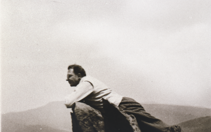 Ein Mann lehnt liegend über eine Bergspitze, im Hintergrund sieht man ein Bergpanorama