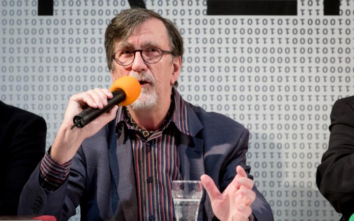 Bruno Latour during the press conference for the exhibition "Reset Modernity" 2016. He holds a microphone in his right hand and makes an open, explanatory gesture with his left hand. A glass of water is in front of him.