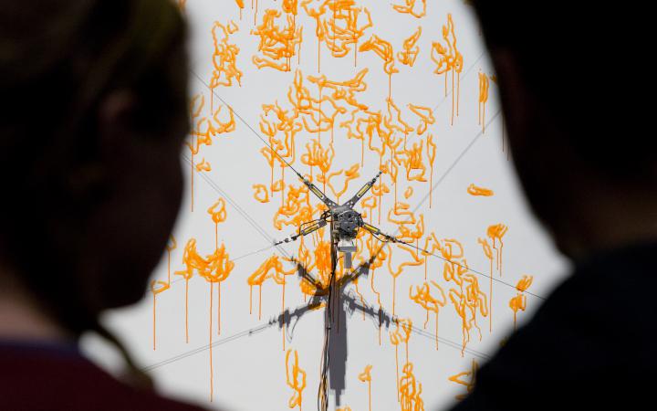  Two silhouettes looking at the computer-controlled robot painting with a yellow paint on a canvas.