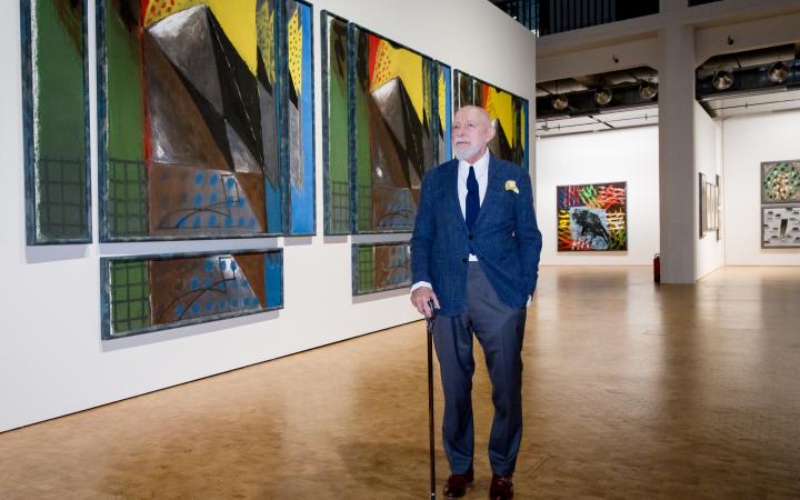 Markus Lüpertz steht im Anzug vor einem bunten Bild in der Ausstellung