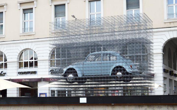 A light blue VW Beetle in a latticework scaffold stands on a pedestal on Karlsruhe's market square.