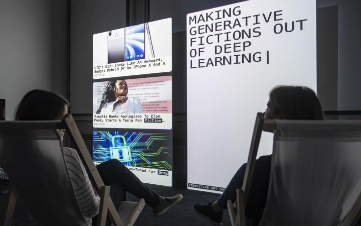 Two people sit in deck chairs in front of two large screens. On the right screen is written "Making generative fictions out of deep learning".