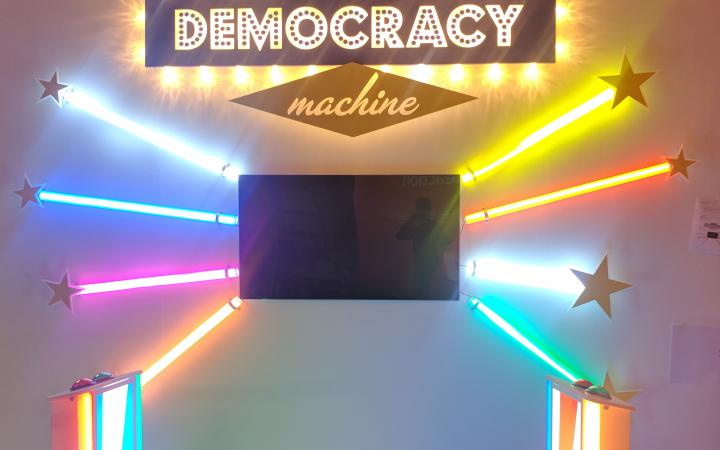 Frontalansicht der The Democracy Machine!. In der Mitte einer Wand ist ein Fernseher installiert darum herum sind je drei bunte Farbröhren wie Sonnenstrahlen angeordnet. Davor stehen zwei Podeste mit Knöpfen.