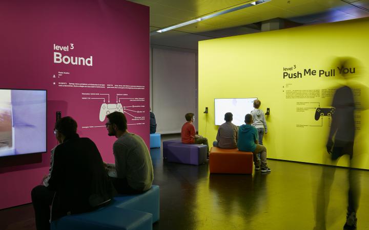 Links im Vordergrund sitzen auf einer Sitzbank zwei Besucher vor einem Bildschirm, der an einer magentafarbenen Wand befestigt ist. Rechts etwas im Hintergrund sitzen mehrere junge Besucher und Besucherinnen vor einer gelben Wand mit Bildschirm. 