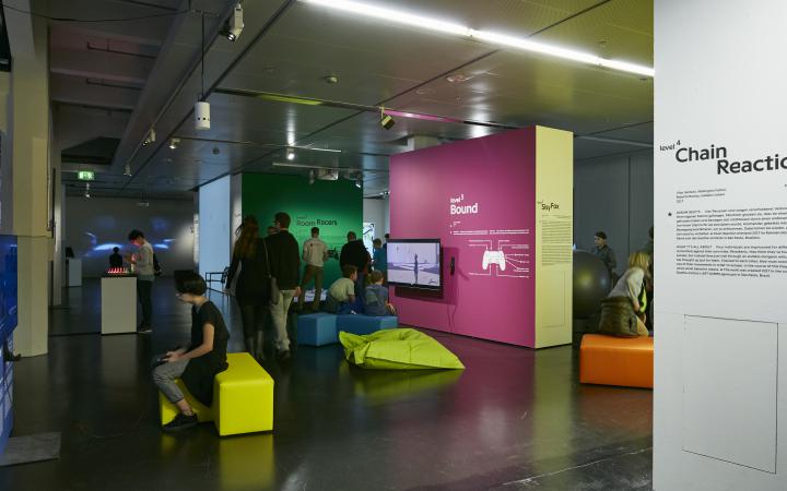 View into the exhibition room with several colourful partitions and exhibits. Numerous visitors standing or sitting on colourful seats.
