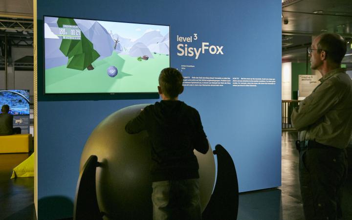 A young visitor in front of a game console consisting of a large ball on a rack. Behind a screen on a blue wall. Another visitor is standing next to it. 