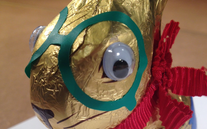 A golden chocolate Easter bunny looks into the camera with its wobbly eyes glued on. Beside the eyes a green sticker similar to glasses is glued on the packaging of the Easter bunny.