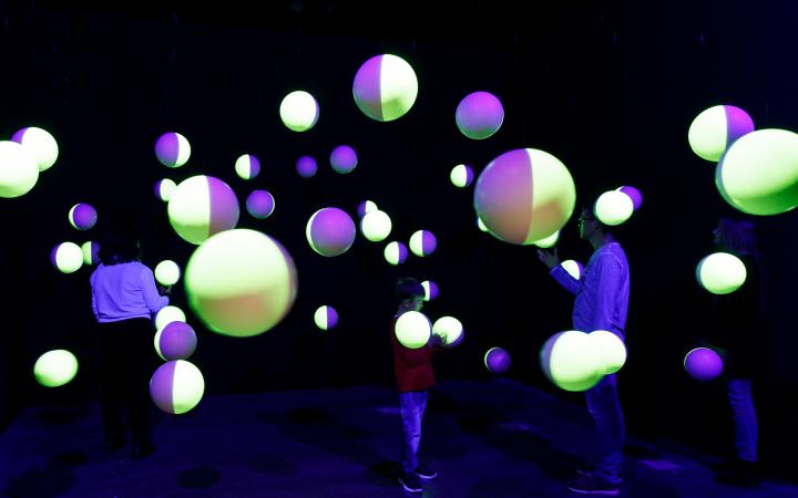 In einem dunklen Raum hängen halbseitig-fluoreszierende Styroporkugeln von der Decke mit denen drei Besucher interagieren