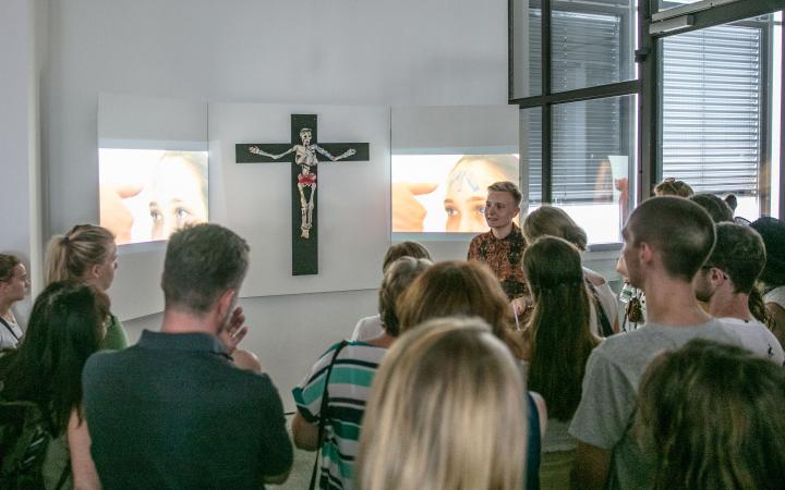 An der Wand hängt ein Kreuz mit einem Gerippe, als Triptychon werden daneben zwei Videos gezeigt. Vor dem Kunstwerk steht eine Menschenmenge.