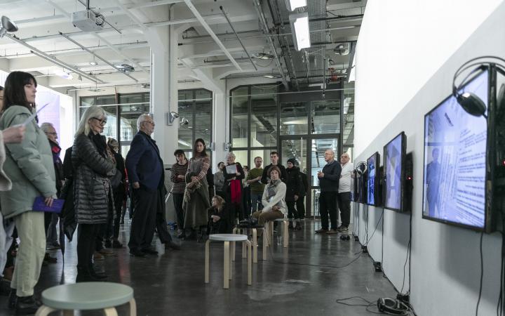 The photo shows the museum balcony of the ZKM with the exhibition of the conference. On the right there are three screens with headphones and opposite are many people.