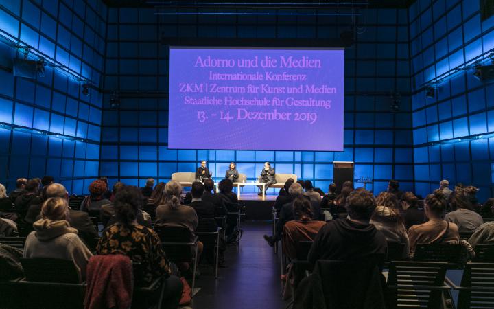 In the darkened media theatre, the poster of the Adorno and the media conference illuminate. Johan Hartel, Loudmila Voropai and Peter Weibel sit on the podium from left to right.