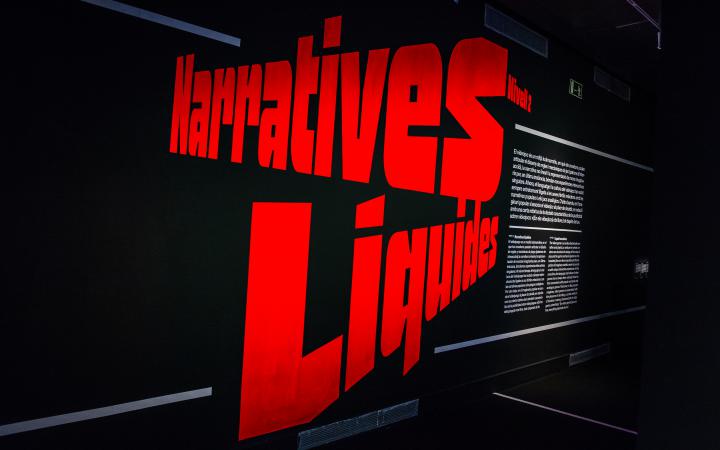 Der Schriftzug »Narratives Liquides« ist in großen roten Buchstaben in schräglage zu sehen. Rechts beginnt der Ausstellungseingang.