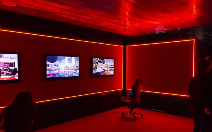 The photo shows a red illuminated room with elegant large office chairs. Three monitors are mounted on the wall, with the chairs in front of them.