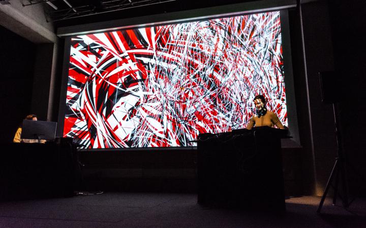 A visual-sound performance will be shown. Two women are standing at the bottom corners of the large screen at their sound controllers while in the background a colour strip of red, black and white merge into one another.