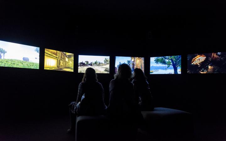 The photo shows three women sitting in a dark room, with only their silhouette visible. In a semicircle there is a row of screens surrounding the women.