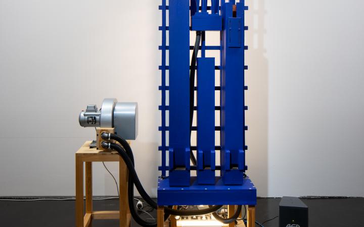 On display is a rectangular, machine-like installation standing on a stool. To the left of the installation there is also a small stool on which an object lies. 