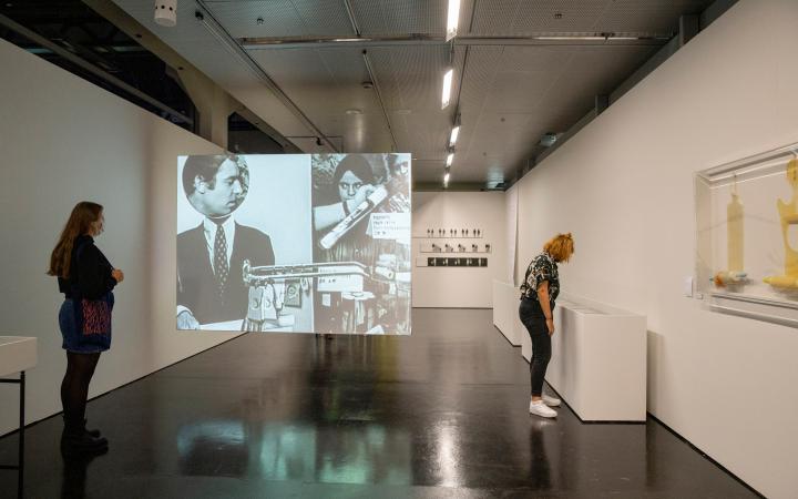 Two young women in the exhibition space. One is standing on the left wall and the other is looking at the right wall in showcases. A screen is stretched between them, on which a film clip with people can be seen.