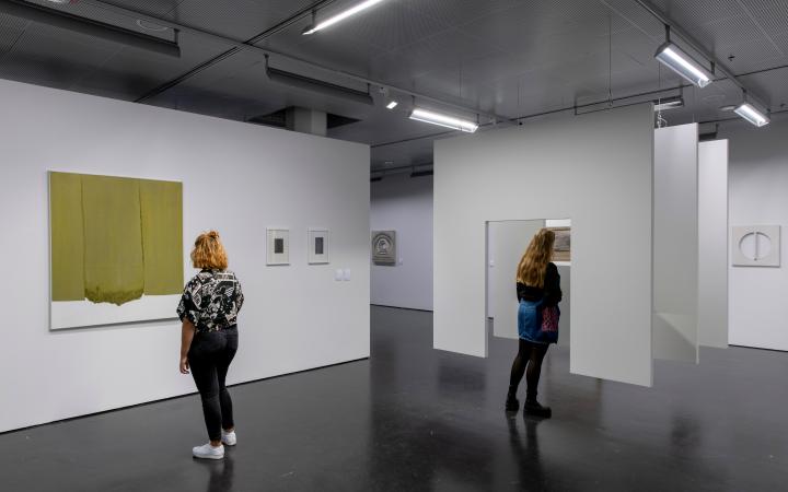 Two women in the exhibition room. On the left a woman is looking at a monochrome painting on the wall. In the middle, a woman is standing in front of a hanging construction consisting of three successive surfaces, with a passageway in between.