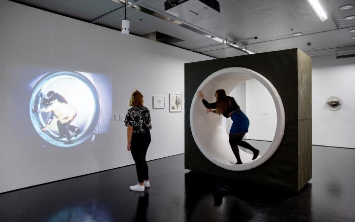 Zwei junge Frauen sind im Ausstellungsraum. Eine betrachtet eine Projektion eines künstlerischen Films an einer Wand und die andere befindet sich in einer Lauftolle.