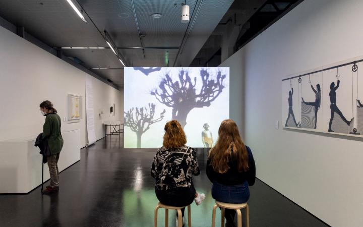 Three young women in the exhibition space. Two sit on stools in front of a film screen on which bare trees can be seen. An abstract drawing hangs on the wall to the right. On the left wall are boxes into which the third woman looks.