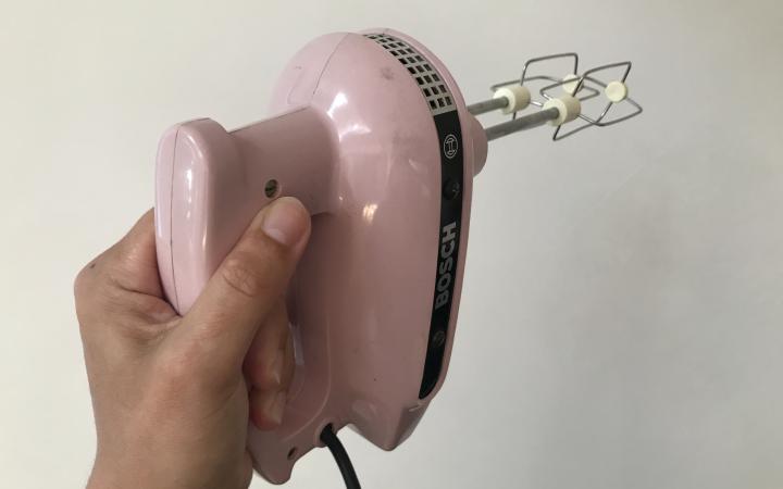 A pink hand mixer is lifted horizontally in front of a white wall.